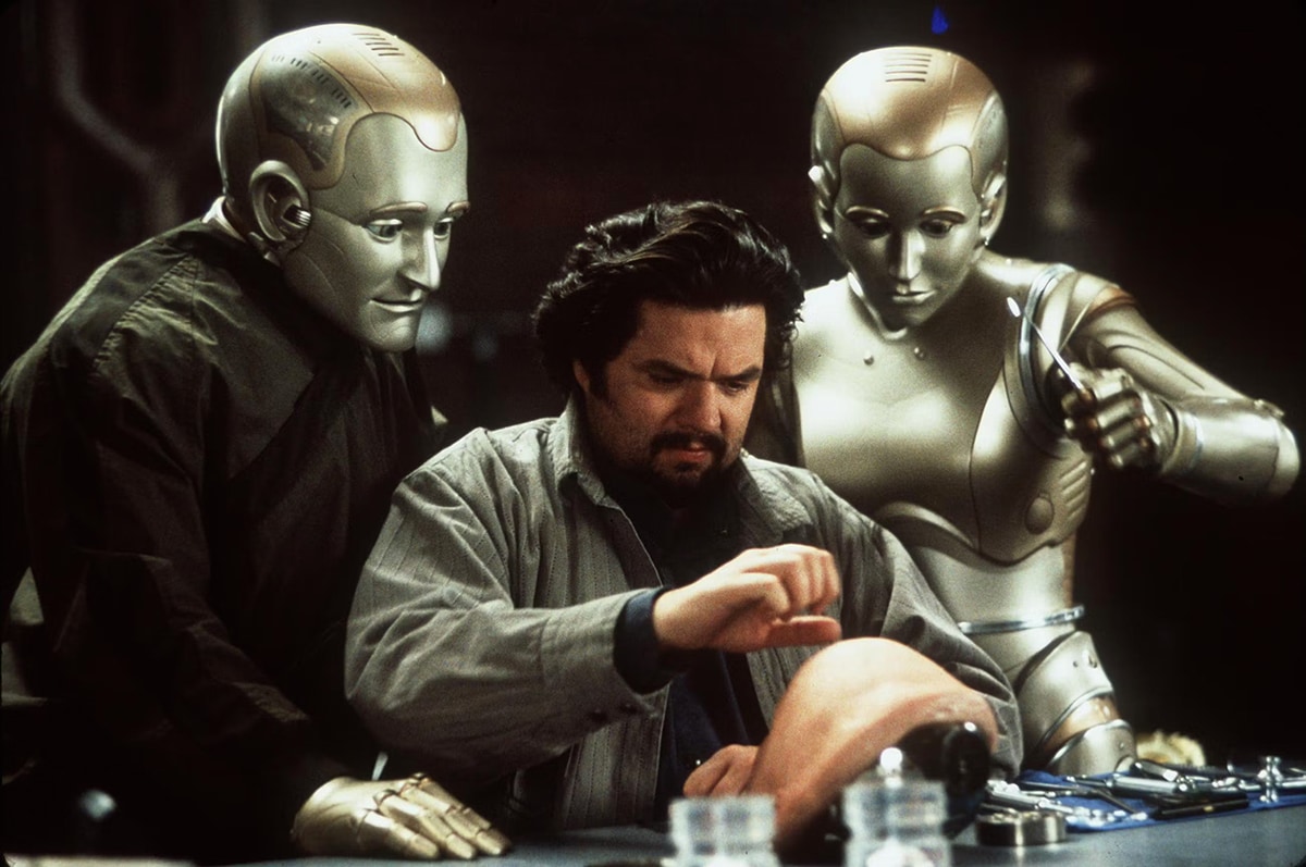 Scene from Bicentennial Man: a man works on a laptop while two golden humanoid robots watch intently over his shoulders